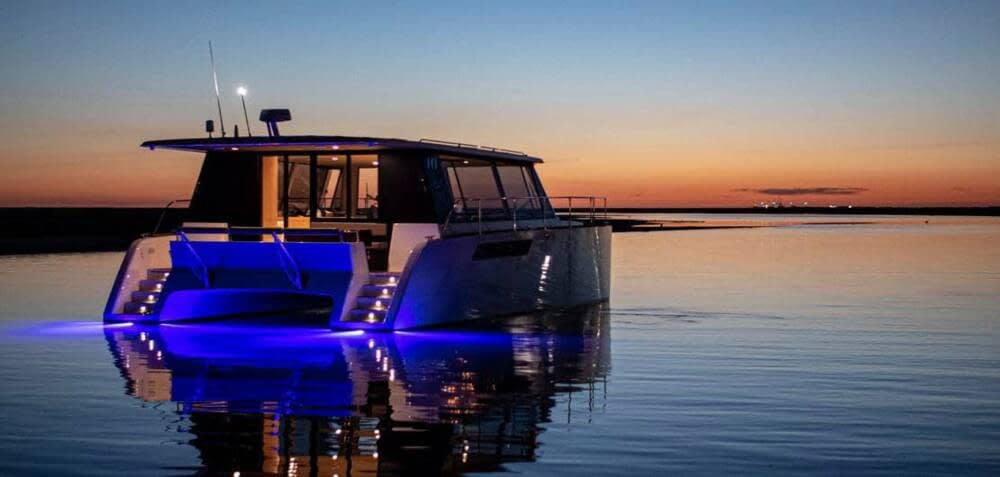 The catamaran at dusk with its deck lights on, low sun on the water